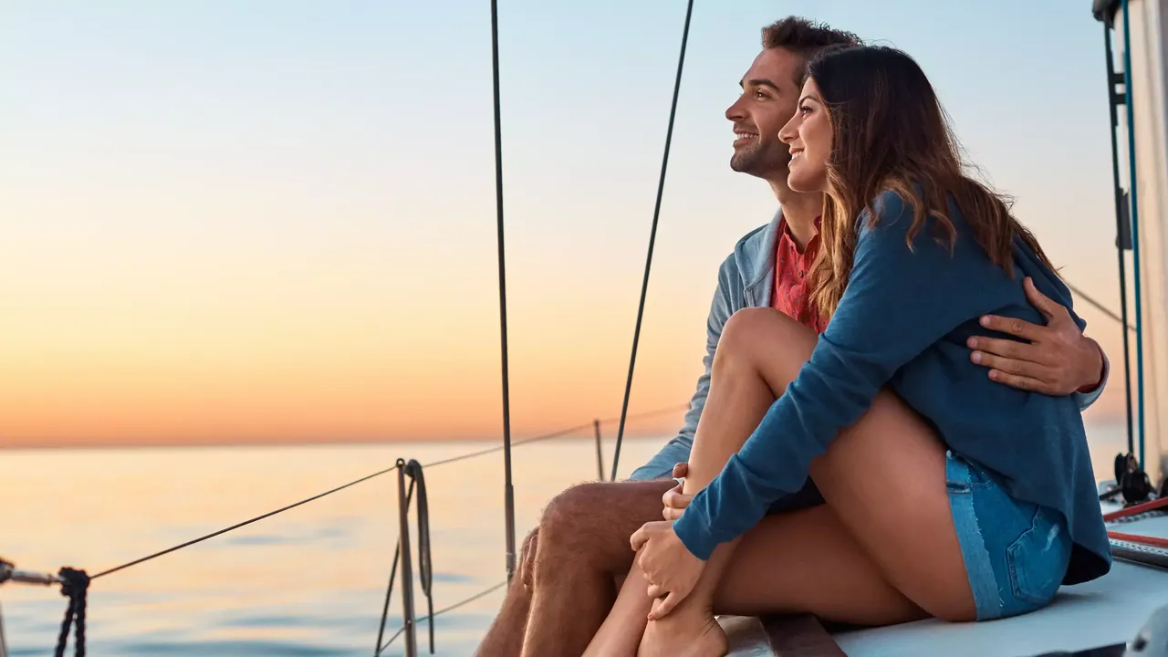 Ein dunkelhaariges Paar in Jeansshorts und Jeanshemden sitzt auf einer Jacht und genießt den Sonnenuntergang über dem Meer.