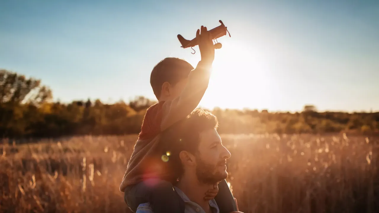 Mann in blauen Hemd trägt ein Kind auf seinen Schultern, welches mit einem Spielzeug-Flugzeug spielt. Im Hintergrund geht die Sonne unter.