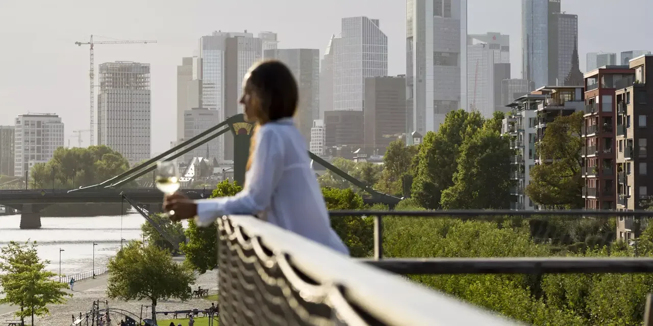 Außenansicht: Eine Frau steht auf einem Balkon der hausInvest Immobilie One Forty West in Frankfurt am Main und schaut auf die Frankfutert Skyline