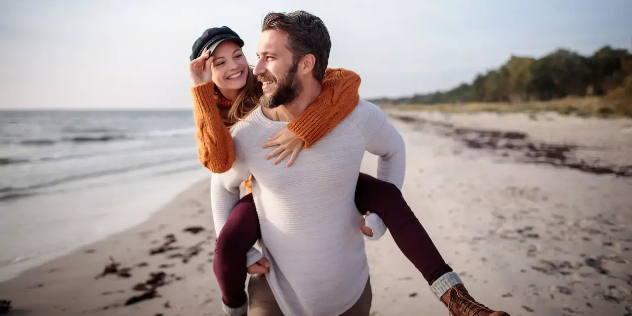 Ein Mann mittleren Alters in einem weißen Hemd trägt seine lächelnde Freundin in einem orangefarbenen Pullover und einer schwarzen Baskenmütze auf dem Rücken, während er lächelnd am Strand spazieren geht.
