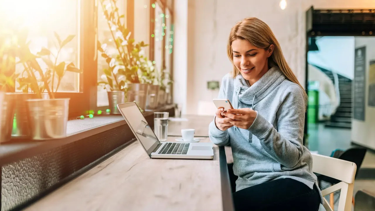 Eine Frau mit hellen Haaren sitzt lächelnd in einem grauen Kapuzenpulli auf dem Stuhl in einem Open-Space-Office mit ihrem Laptop auf dem Tisch und einer Tasse Kaffee und einer Tasse Wasser, und schreibt auf ihrem Handy.