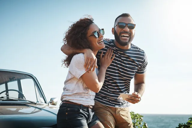 Zwei lächelnde Personen – ein Mann in einem gestreiften T-Shirt und einer Sonnenbrille und eine Frau in einem weißen T-Shirt und einer Sonnenbrille – umarmen sich vor einem Auto stehend; im Hintergrund ist ein Meer zu sehen.