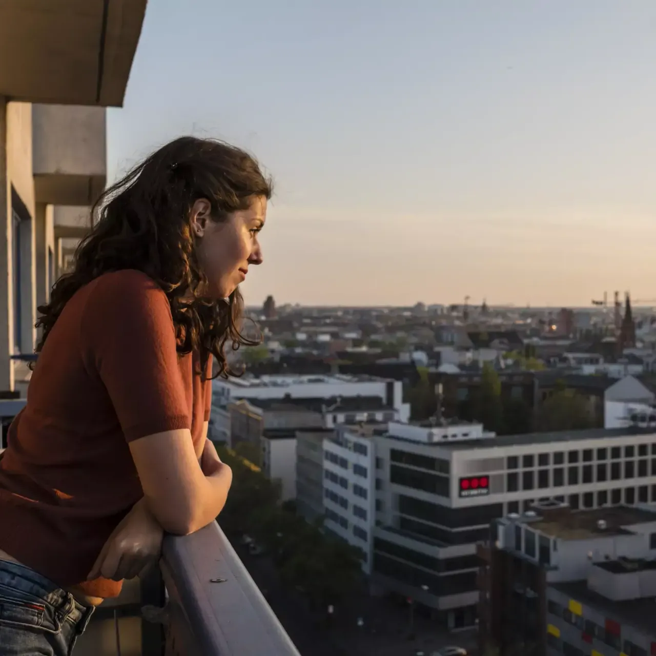 Eine junge Frau steht auf ihrem Balkon angelehnt an die Mauer und schaut auf die Stadt Berlin.