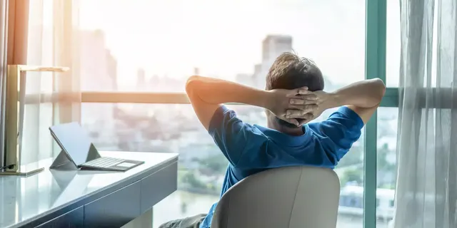 Ein Mann in einem blauen T-Shirt sitzt in seinem Rollstuhl mit dem Rücken zur Kamera und mit seinen Händen hinter dem Kopf und guckt durch Glaswände in die Ferne; auf seinem Tisch ist ein Laptop zu sehen.