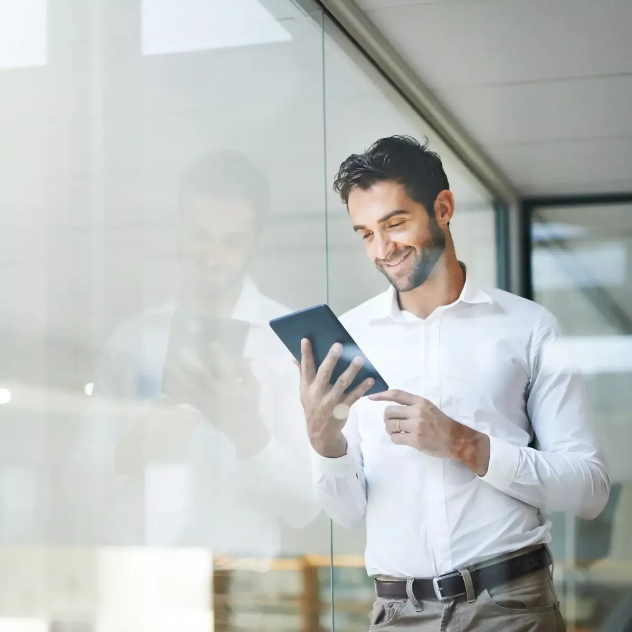 Ein junger Mann mit einem weißen Hemd steht im Büro an einer Glaswand und schaut interessiert auf sein iPad