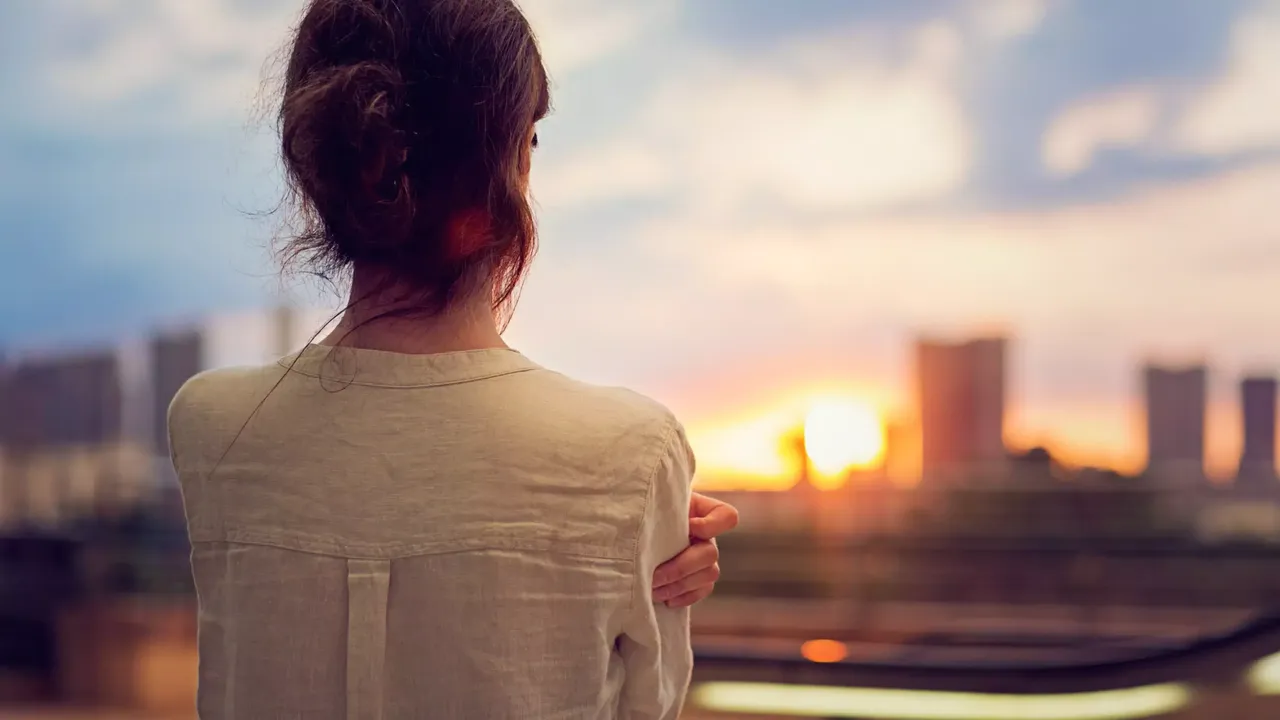 Eine Frau mit einer weißen Bluse und braunen Haaren schaut auf eine Skyline bei Sonnenuntergang.