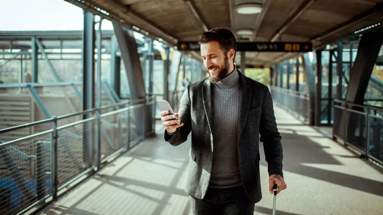 Ein dunkelhaariger bärtiger Mann in einem grauen Pullover und einer grauen Jacke steht mit seinem Koffer auf dem Bahnsteig und schaut auf sein weißfarbiges Handy.