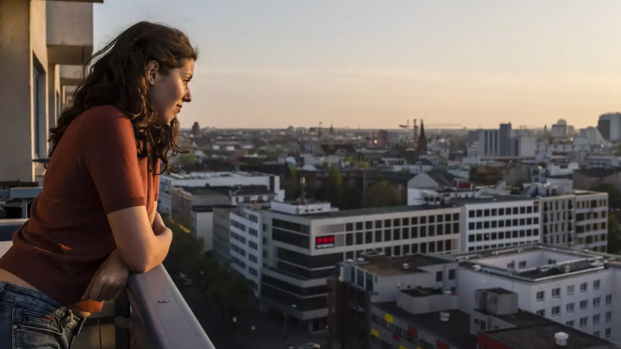 Eine junge Frau steht auf ihrem Balkon angelehnt an die Mauer und schaut auf die Stadt Berlin.