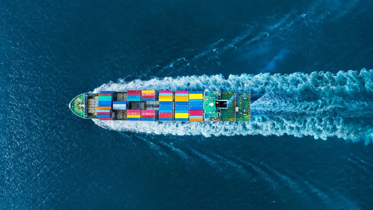 Luftaufnahme eines Containerschiffs mit bunten Frachtcontainern, das auf tiefblauem Meer fährt und eine breite Bugwelle hinterlässt.