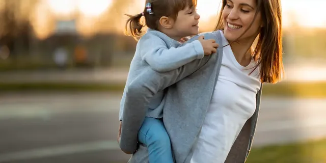 Die Mutter in einem weißen T-Shirt auf der Straße sieht ihre kleine Tochter in einem hellblauen Cardigan an, welche auf ihrem Rücken sitzt und lächelt.