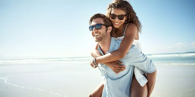 Ein junger Mann in einem hellblauen Hemd und einer Sonnenbrille trägt seine Freundin in einem weißen T-Shirt und einer Sonnenbrille auf dem Rücken am sonnigen Strand.