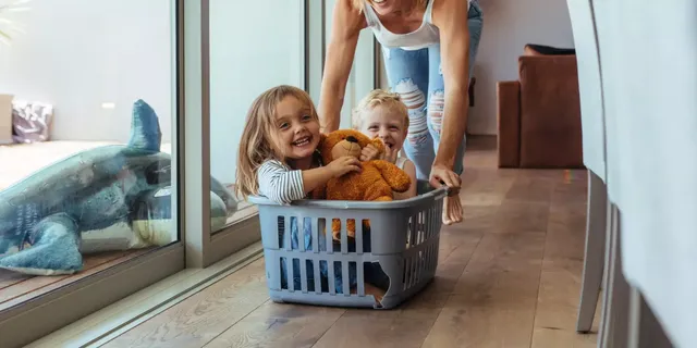 Die lächelnde Mutter mit blonden Haaren in einem weißen T-Shirt und in den Jeans spielt zu Hause mit ihren kleinen Kindern – einem blonden Mädchen mit dem braunen Teddy-Bär und einem blonden Junge, – welche in einem Wäschekorb sitzen und lächeln.