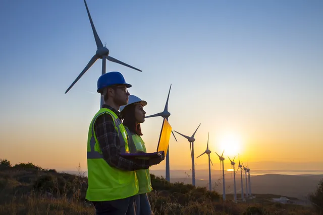 klimaVest: Zwei Arbeiter mit Warnwesten
  und Helmen sowie einem Laptop stehen vor einem Windpark. Im Hintergrund geht
  die Sonne unter.