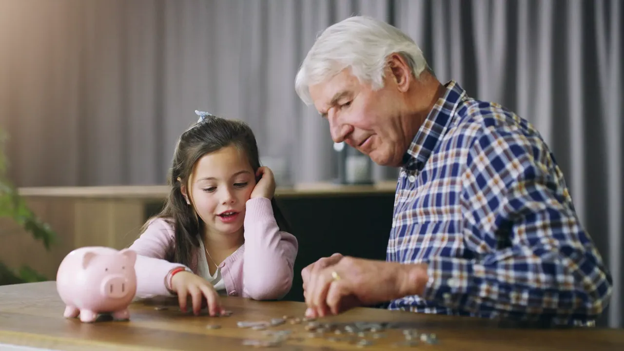 Ein Großvater mit weißen Haaren im karierten Hemd sitzt mit seiner dunkelhaarigen Enkelin in einem rosa Cardigan am Tisch und hilft ihr, die Münzen aus ihrem rosa Sparschwein zu zählen.
