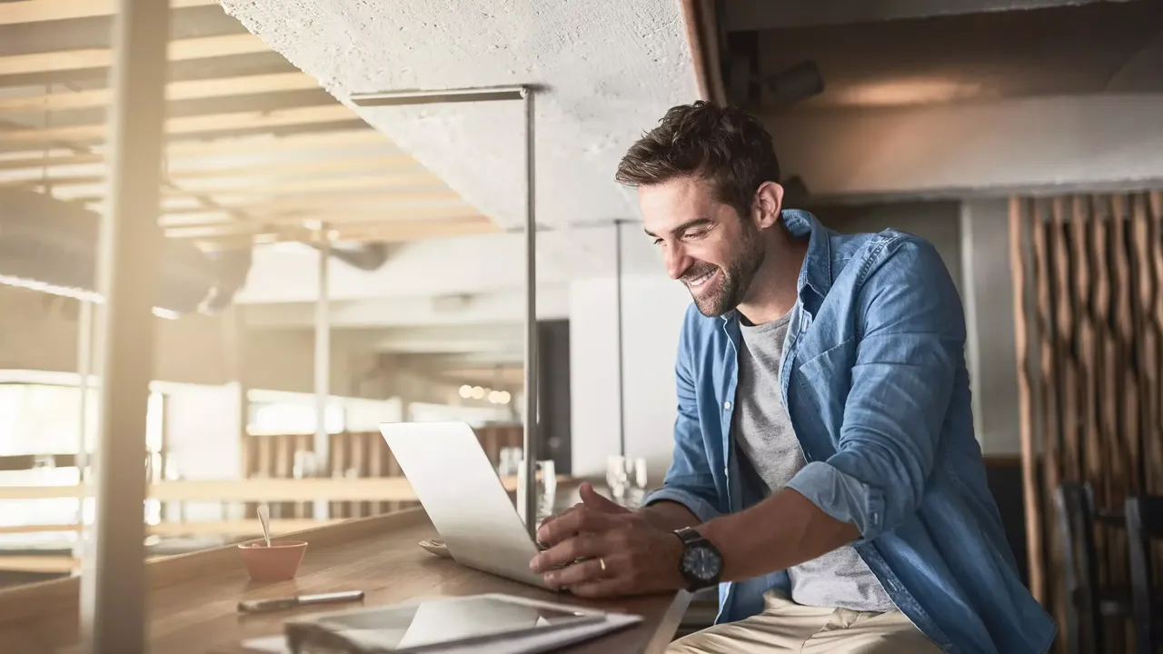 Ein junger dunkelhaariger bärtiger Mann sitzt auf dem zweiten Stock des sonnigen Open-Space-Büros in einem weißen T-Shirt, einem Denim-Hemd und in einer khakifarbigen Hose und arbeitet am Laptop, welches auf dem Holztisch neben einem Tablet und Dokumenten sich befindet.