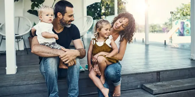 Eine Familie – ein bärtiger Mann in einem schwarzen T-Shirt und Jeans, ein kleiner Junge in einem weißen T-Shirt und Shorts mit einem Pflanzenmuster, ein kleines Mädchen in einem khakifarbigen Kleid und eine Frau in einem weißen T-Shirt und Jeans – sitzen draußen auf der graufarbigen Holzveranda ihres Hauses und lächeln; im Hintergrund sind zwei weiße Stühle und eine Pflanze in einem Topf zu sehen.