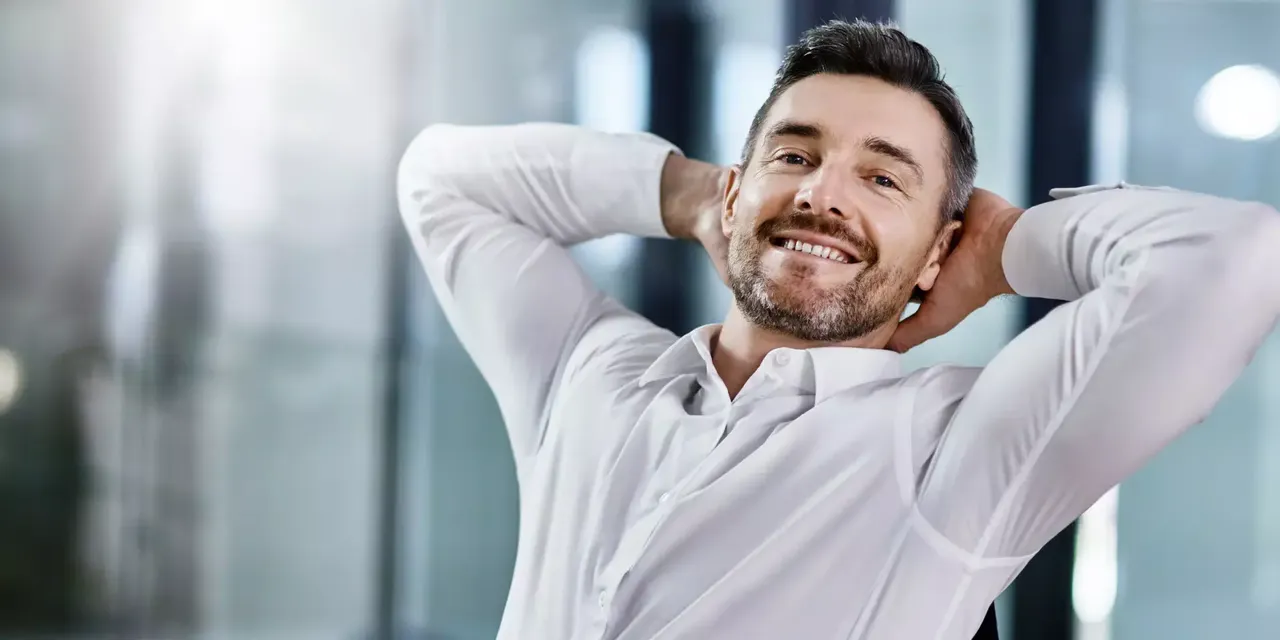 Ein lächelnder Mann mittleren Alters in einem weißen Hemd sitzt im Bureau mit seinen Händen hinter dem Kopf.