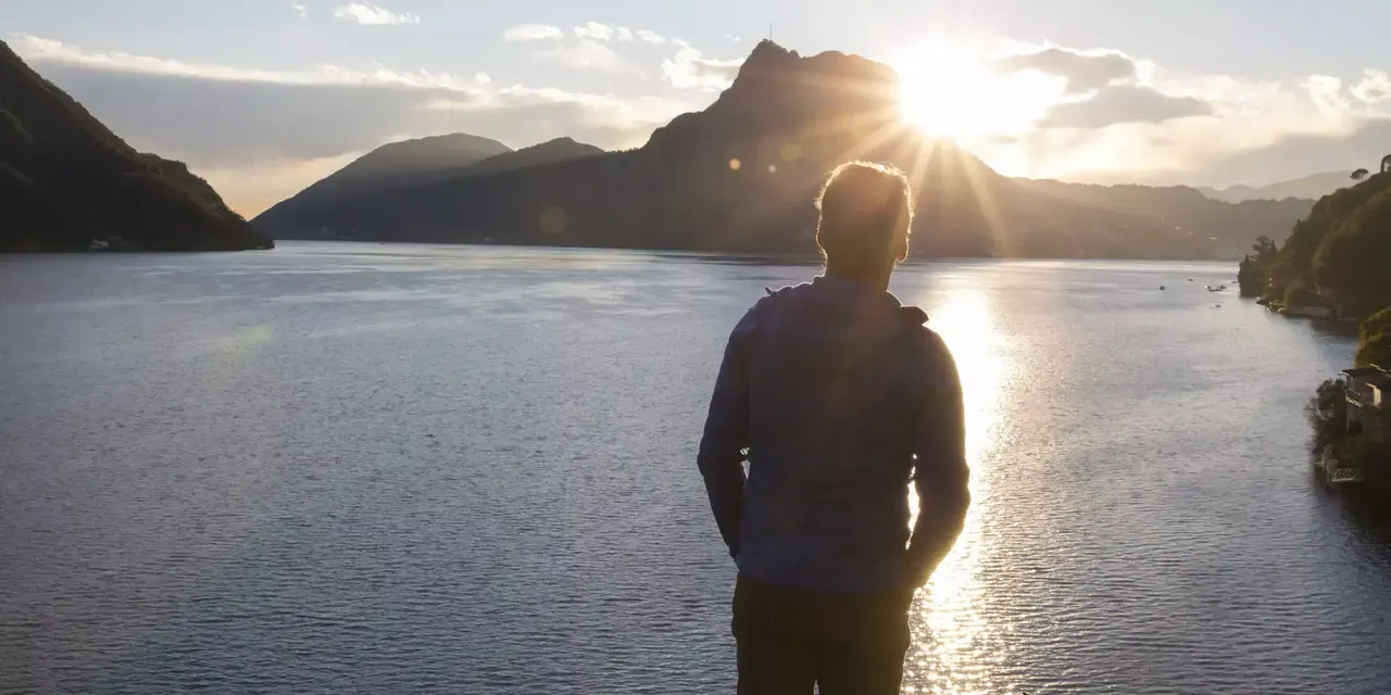klimaVest: Mann steht mit Händen in den Hosentaschen am Ufer des Meeres und guckt sich den Sonnenuntergang an.
